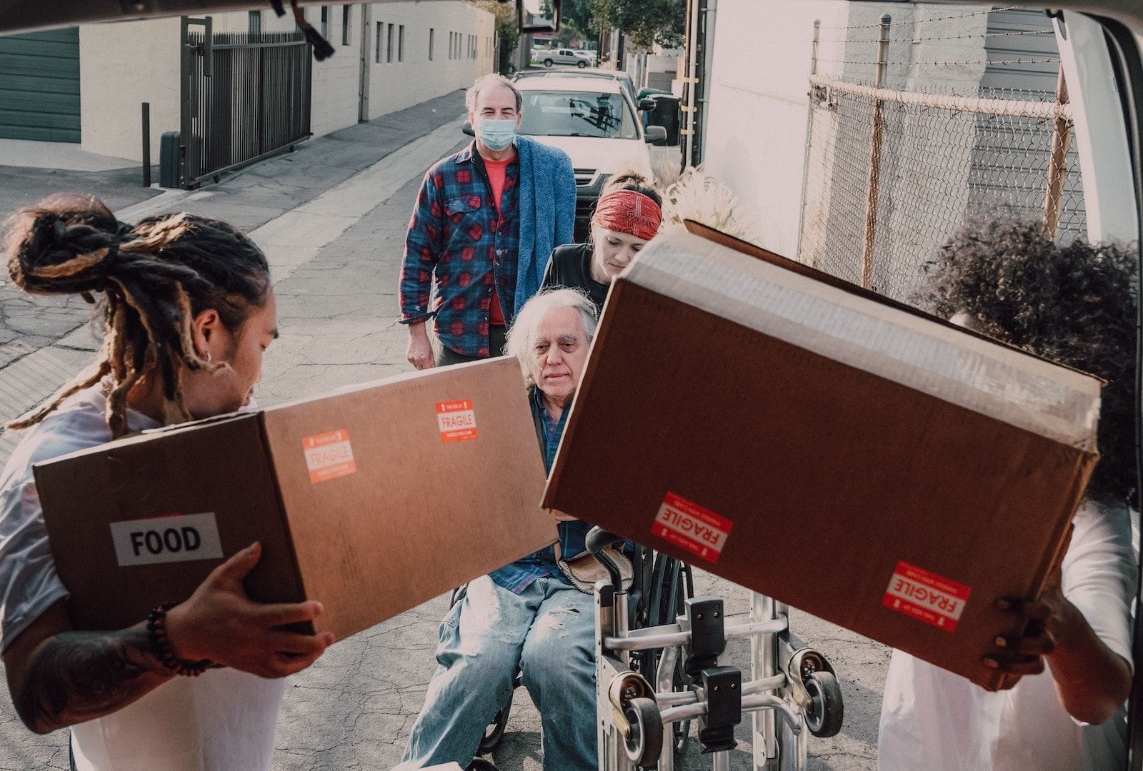 A group of volunteers unloading and handing boxes of food to an elderly individual in aa wheelchair.